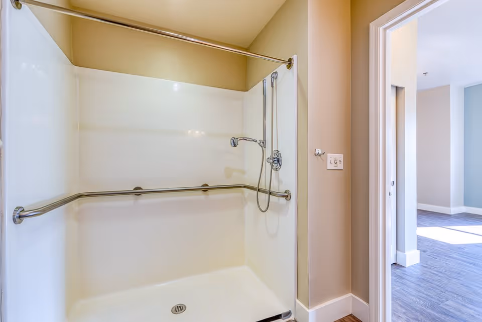 A clean, accessible shower area with beige walls, a handheld showerhead, and a horizontal grab bar. The shower has a white surround and a curtain rod above. To the right, there is an open doorway leading to a room with light blue and beige walls and wood flooring.