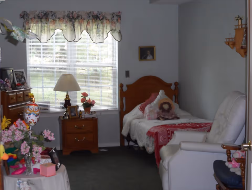 Small furnished bedroom with a single wooden bed, nightstand beneath a window, armchair, and floral decorations.