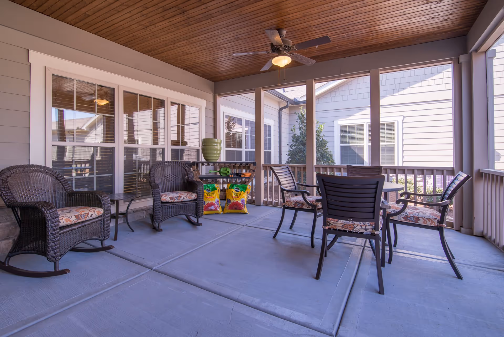 A covered outdoor patio area with a wooden ceiling and ceiling fan. The patio features two wicker rocking chairs with patterned cushions, a small side table, and a round table with four chairs that have matching patterned cushions. The patio is enclosed with railings and overlooks the exterior of the building with windows and siding visible.
