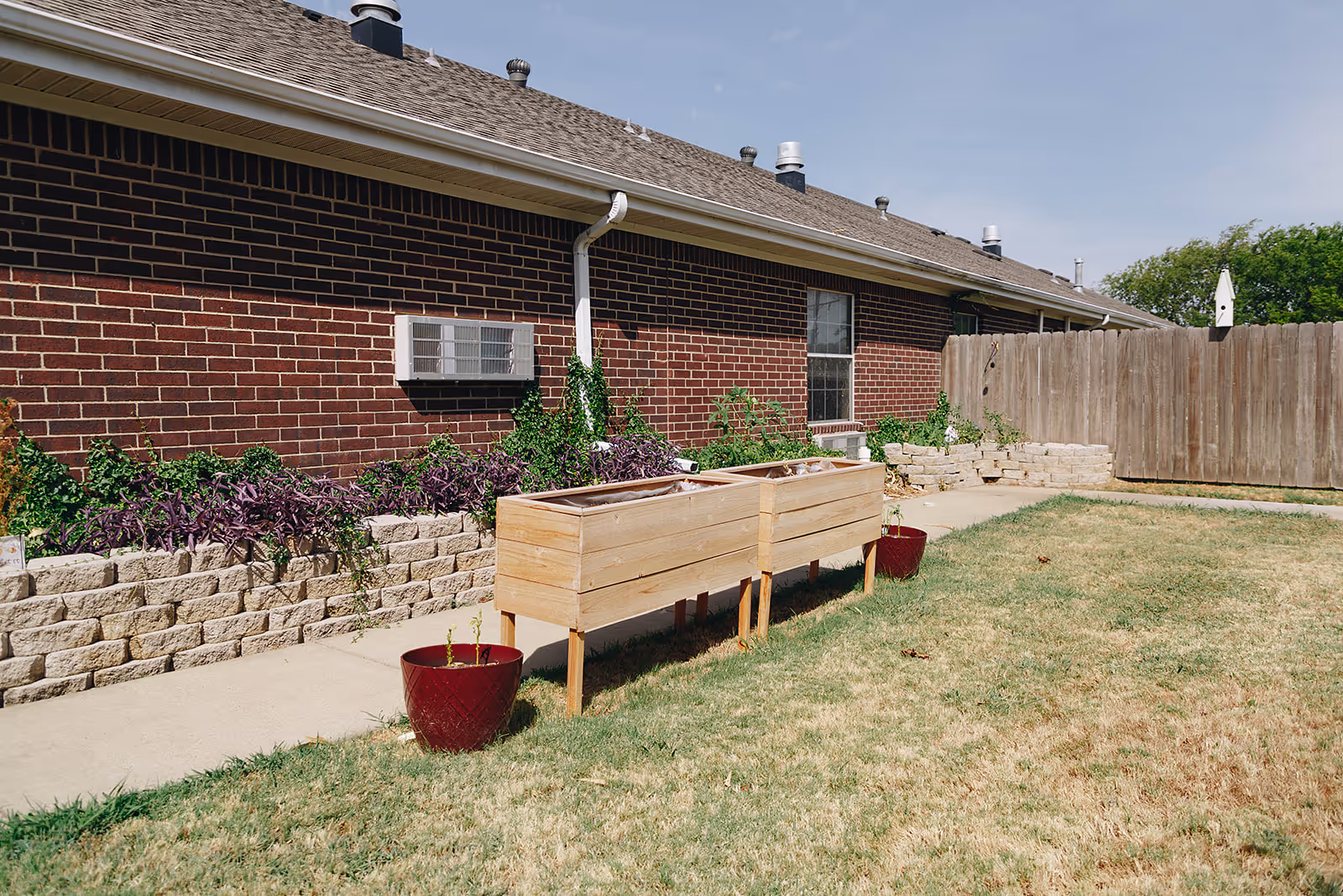 Outdoor garden area with a brick building wall, two wooden raised garden beds, two red plant pots, and a small stone retaining wall with various plants growing. The area is fenced with a wooden fence and has a grassy lawn under a clear sky.