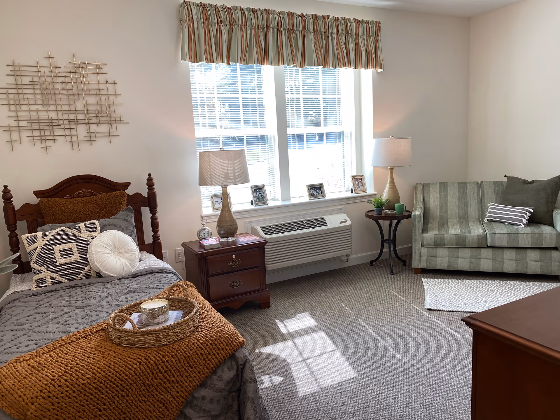 A cozy senior living bedroom with a single wooden bed adorned with multiple pillows and a knitted throw blanket. Next to the bed is a wooden nightstand with a lamp, clock, and framed photos. A large window with blinds and a striped valance lets in natural light. Across from the bed is a green striped loveseat with pillows and a small round side table with a lamp and plant. The room has neutral walls and carpeted flooring.
