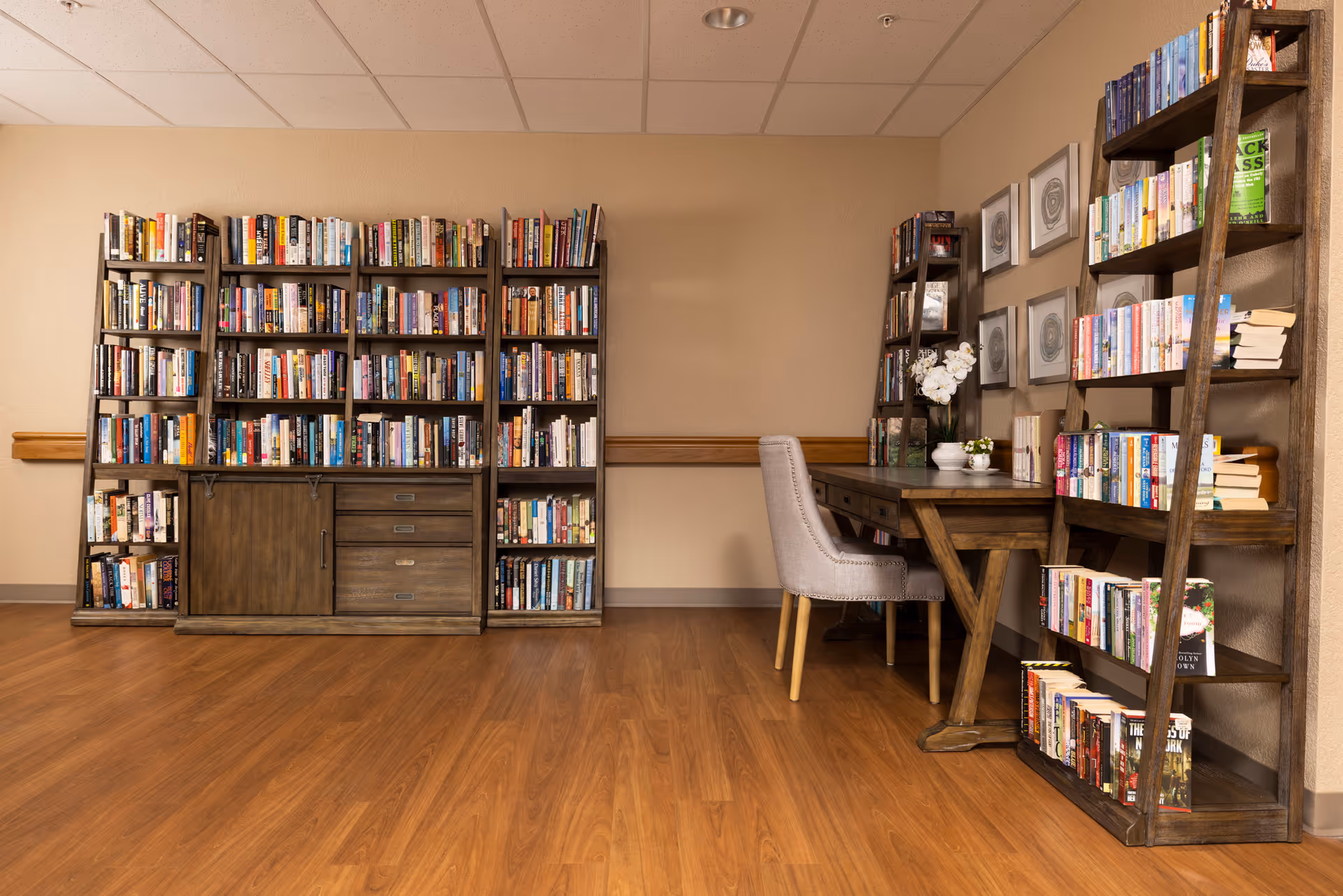 A cozy reading area in a senior care facility featuring wooden bookshelves filled with books, a wooden desk with a cushioned chair, and decorative framed artwork on the wall. The floor is wood, and the walls are beige.