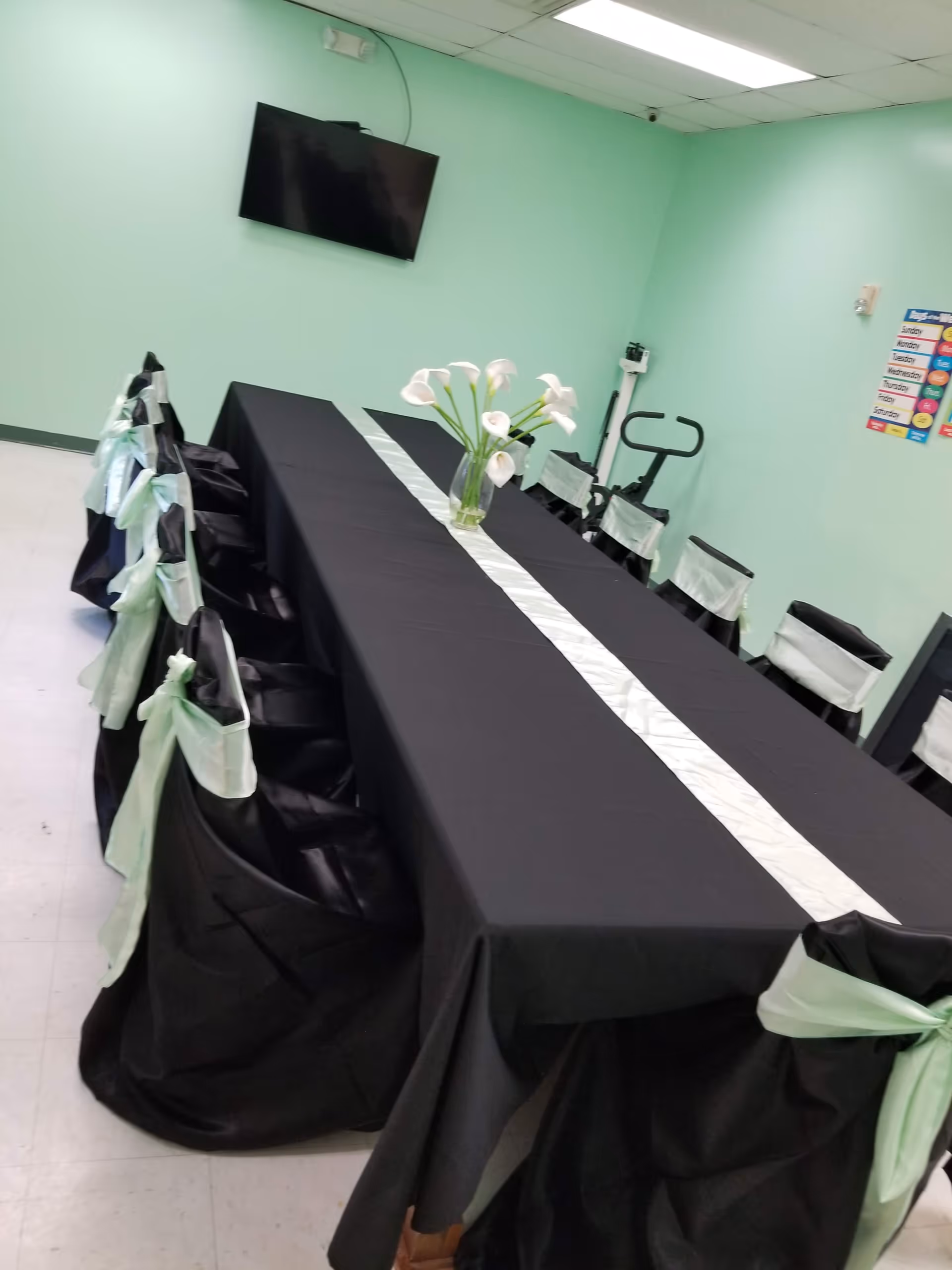 A long rectangular table covered with a black tablecloth and a white table runner, surrounded by chairs draped in black covers with light green bows tied at the back. A vase with white calla lilies is placed in the center of the table. The room has light green walls, a wall-mounted flat screen TV, an exercise bike, and a colorful weekly calendar on the wall.