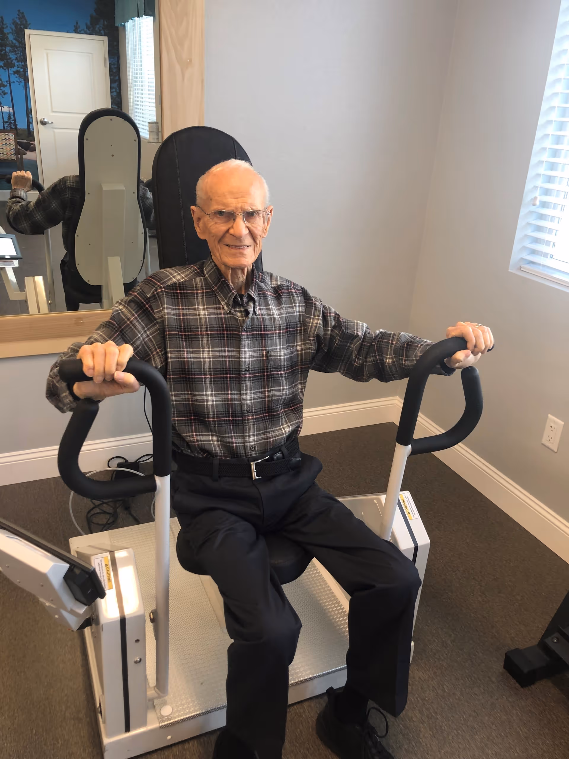 An elderly man wearing glasses and a plaid shirt is seated on a piece of exercise equipment in a room with light gray walls, a window with blinds, and a large mirror reflecting the back of the equipment and part of the room.