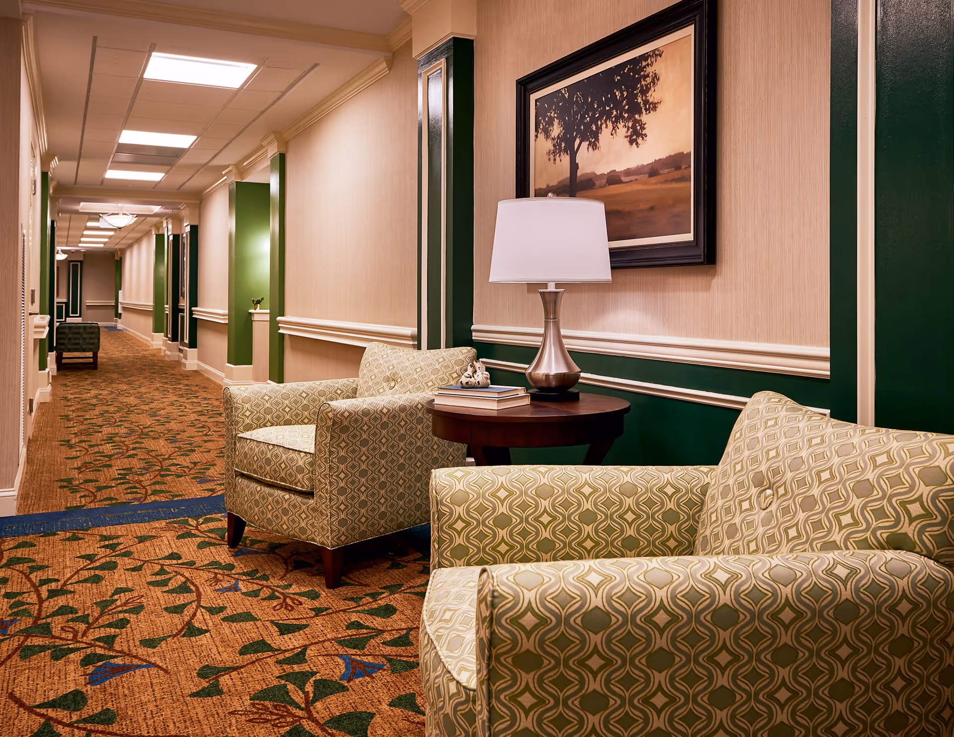 Carpeted hallway seating area with patterned armchairs, a round table and lamp against green-accented walls and framed art.