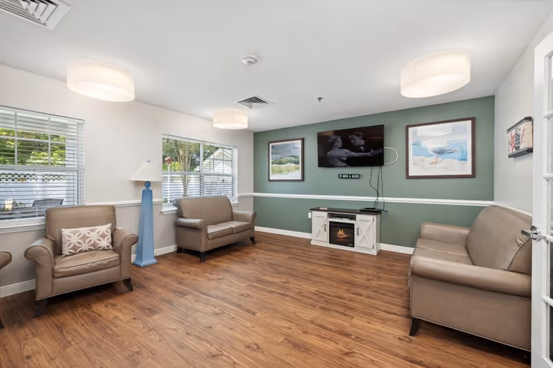 A cozy living room with three brown leather sofas arranged around a small white electric fireplace. The room has wooden flooring, two large windows with white blinds, a blue floor lamp, and three framed pictures on the walls. A flat-screen TV is mounted above the fireplace.