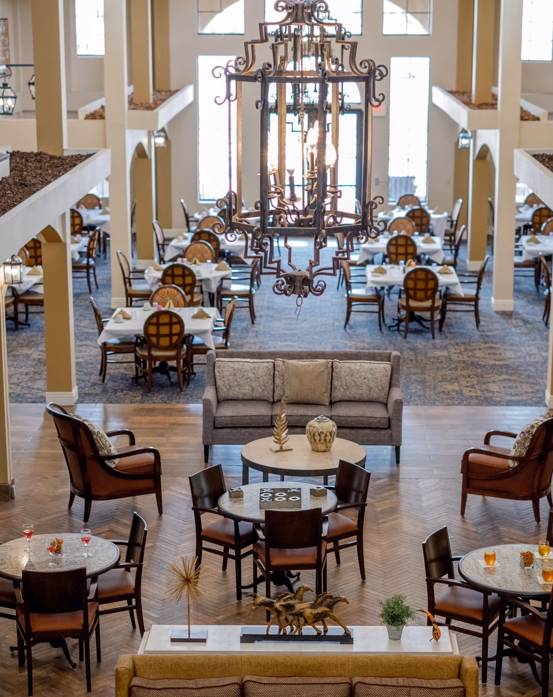 Spacious and elegant dining area in a senior living facility with multiple round and square tables set with napkins and chairs. In the foreground, there is a seating area with a sofa, armchairs, and small tables, one with a tic-tac-toe game. A large ornate chandelier hangs from the ceiling, and tall windows allow natural light to fill the room.