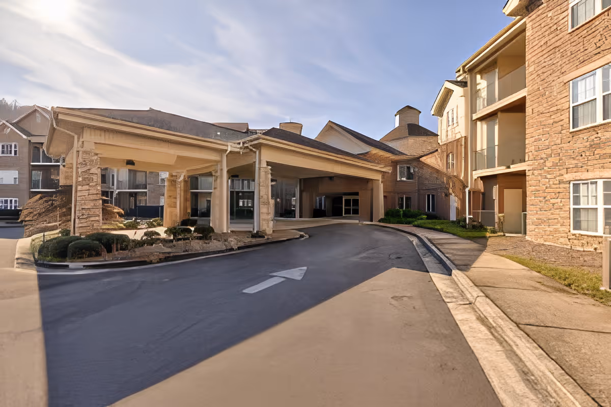 Exterior view of The Haven at Regency Pointe showing the entrance driveway with a covered drop-off area, surrounded by multi-story buildings with stone and brick facades under a partly cloudy sky.