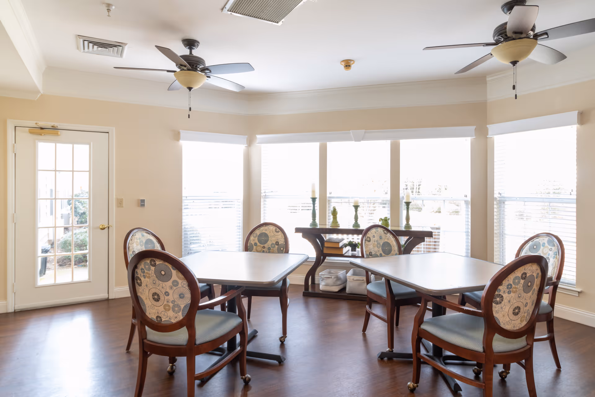 A bright room with large windows and white blinds, featuring two square tables with four upholstered chairs each. The chairs have wooden frames and patterned backs. The room has two ceiling fans with lights, a wooden floor, and a small console table with decorative candles and storage baskets near the windows. A glass door leads outside.