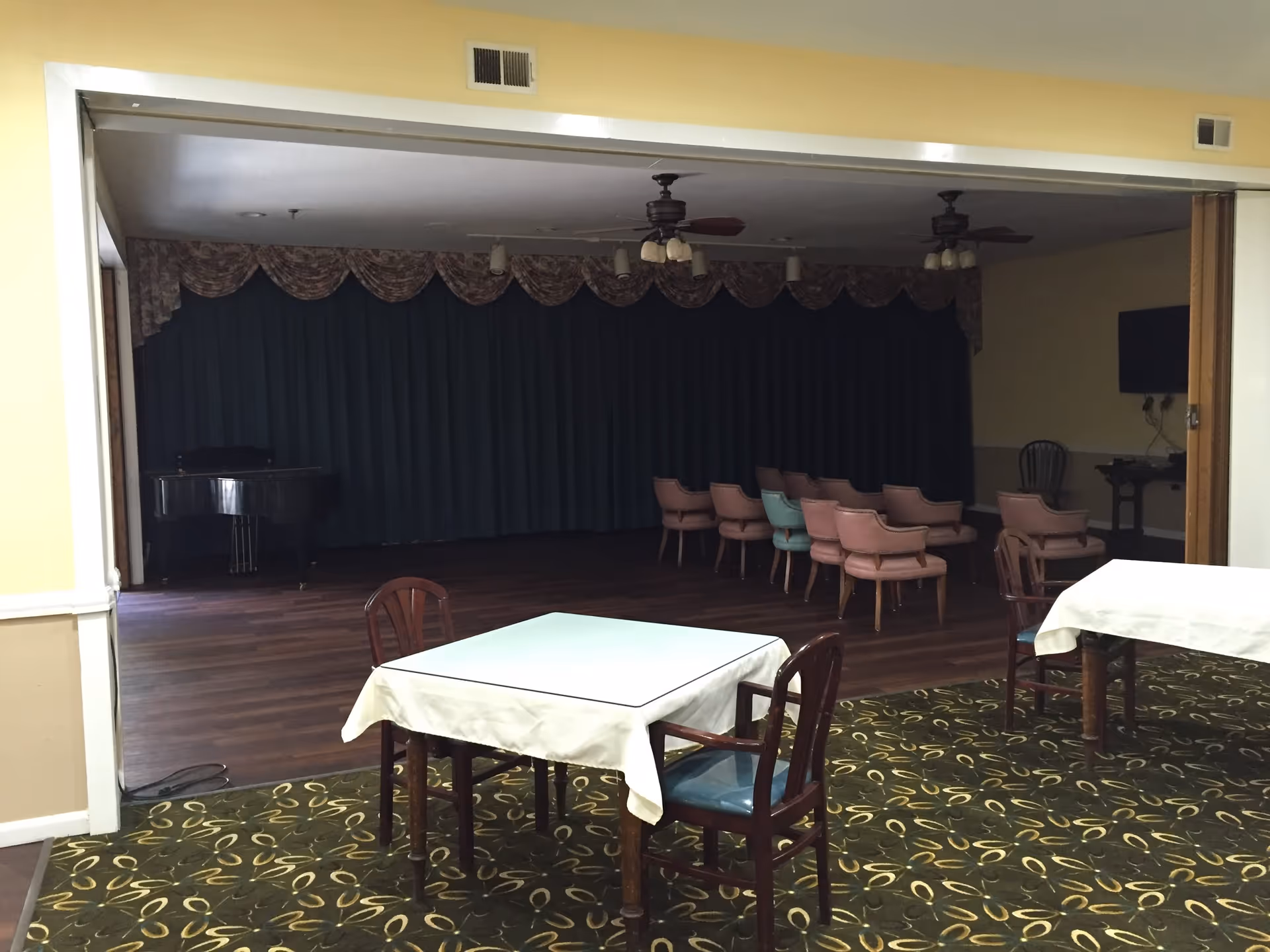 A room with tables covered in white tablecloths and wooden chairs in the foreground, leading to a stage area with a piano and rows of chairs facing the stage. The stage has dark curtains and ceiling fans above.
