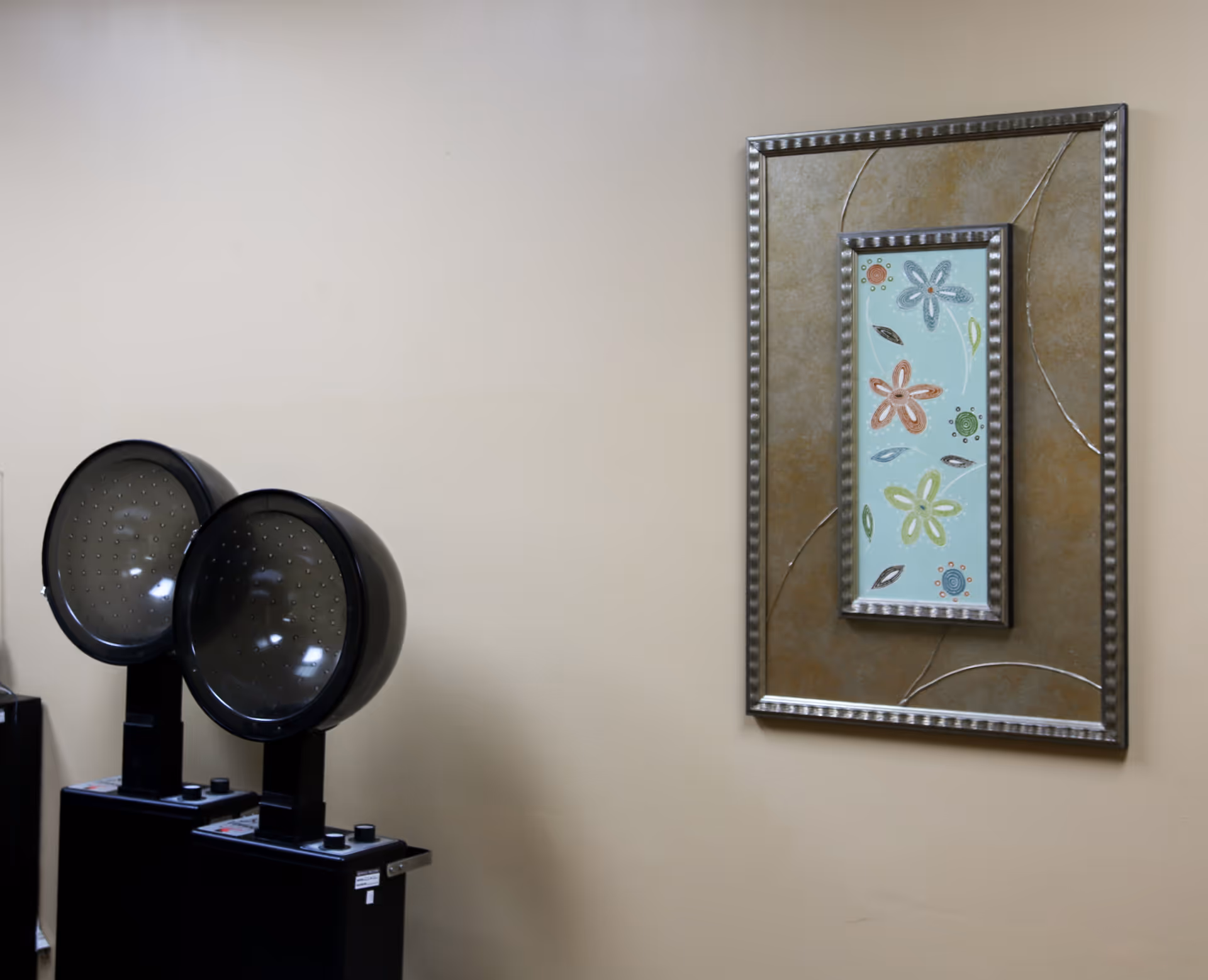 Two black hair dryer chairs positioned against a beige wall with a framed decorative artwork featuring colorful flowers hanging beside them.