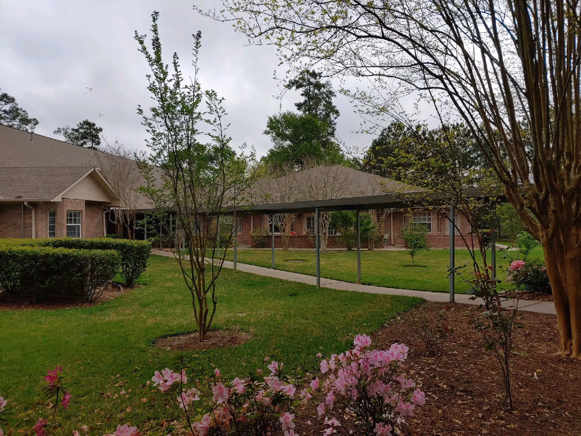 Outdoor view of a senior living facility with a covered walkway, green lawn, trees, bushes, and blooming pink flowers. The building has a brick exterior and a sloped roof under a cloudy sky.