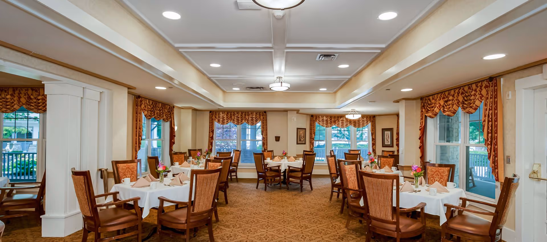 Bright, upscale dining room with round tables set with white linens, wooden chairs, floral centerpieces, and large windows with valances.