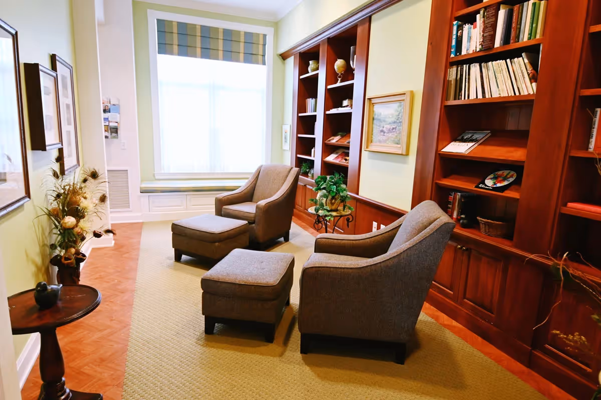 A cozy sitting area with two gray armchairs and matching ottomans on a beige carpet. The room features built-in wooden bookshelves filled with books and decorative items, a small round glass table with a plant, and a large window with a striped valance letting in natural light. The walls are light-colored, and there are framed pictures and a vase with dried flowers on a small wooden table.