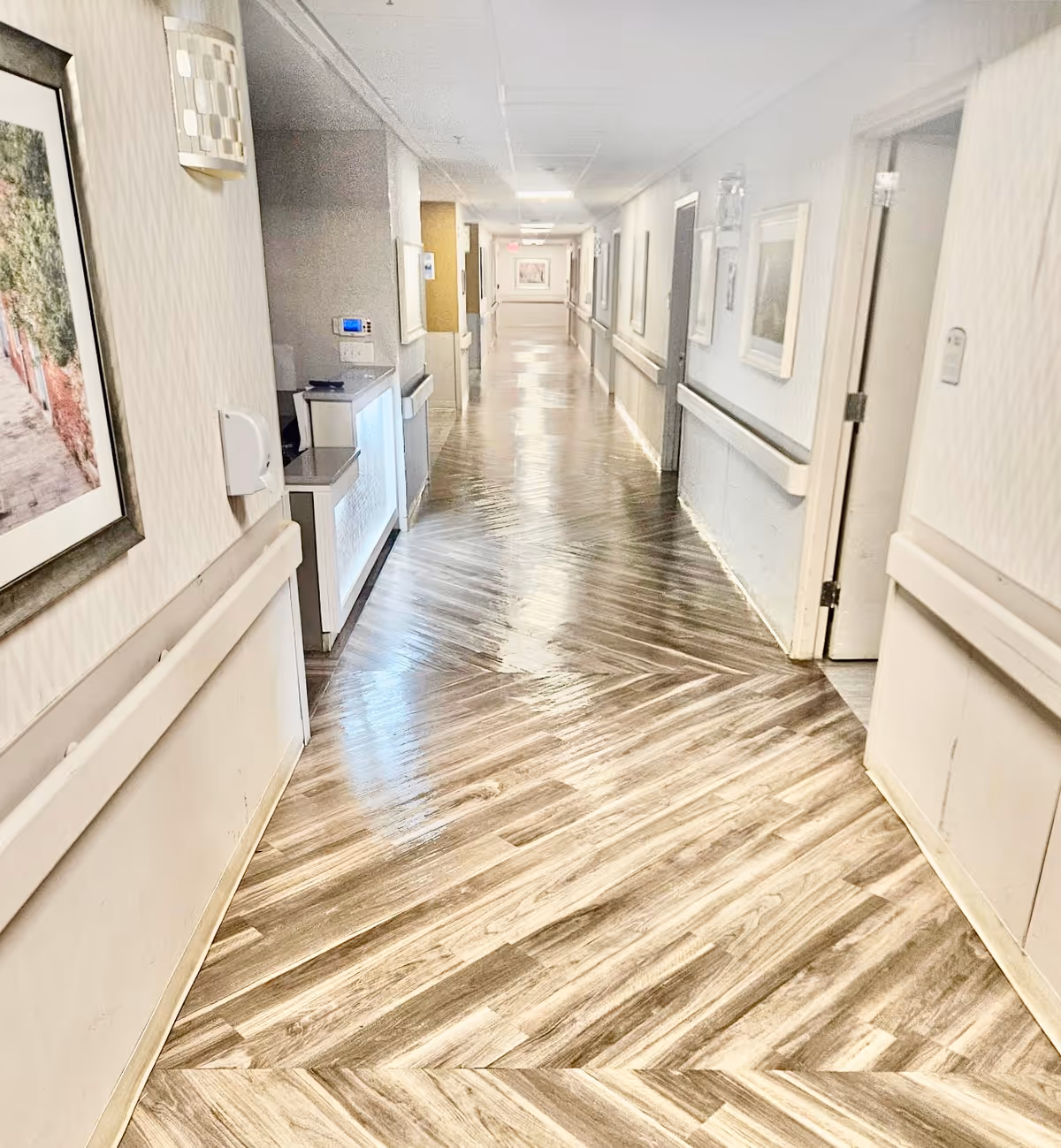 A long, clean hallway in a senior living facility with polished wooden flooring arranged in a herringbone pattern. The walls are light-colored with handrails on both sides and framed artwork hanging along the corridor. Several doorways lead to rooms on the right side, and there is a small counter or desk area on the left side near the foreground.