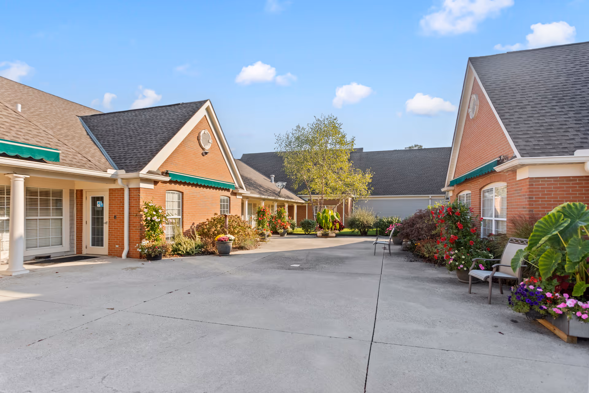 Outdoor courtyard area of a senior living facility with brick buildings on either side, green awnings above windows, potted plants and flowers along the walls, a tree in the center, and a clear blue sky with some clouds.