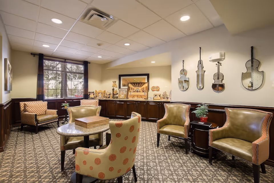 A cozy senior living common area with patterned carpet, a round table with four chairs, two armchairs, and a window with dark curtains. The walls are decorated with violin-shaped mirrors and various framed artworks and sculptures on a wooden cabinet along the back wall.
