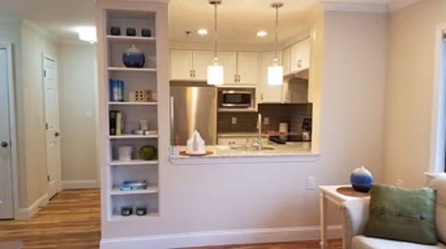 View of a modern kitchen with white cabinets, stainless steel refrigerator, microwave, and a granite countertop with two pendant lights hanging above. To the left is a built-in shelving unit with decorative items, and to the right is part of a living room area with a beige armchair and a small side table holding a blue vase.