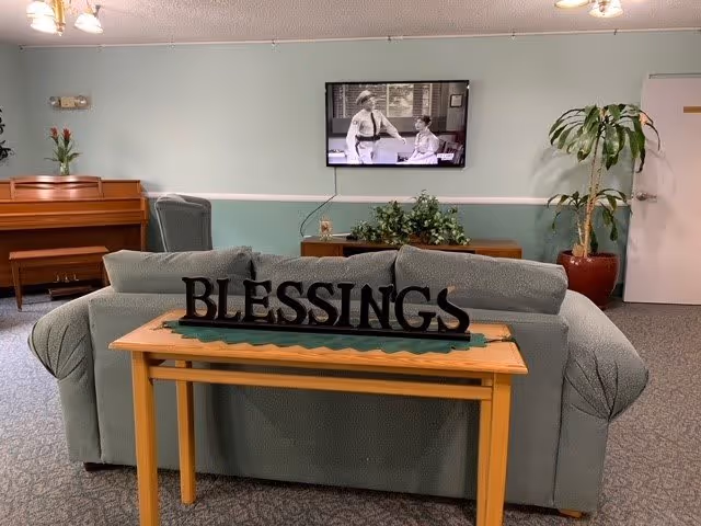 A cozy living room area with a green couch facing a wall-mounted television showing a black and white program. Behind the couch is a wooden table with a decorative sign that reads 'BLESSINGS'. To the left, there is a piano with a vase of flowers on top, and to the right, a large potted plant near a closed door. The walls are painted light green with a white chair rail.