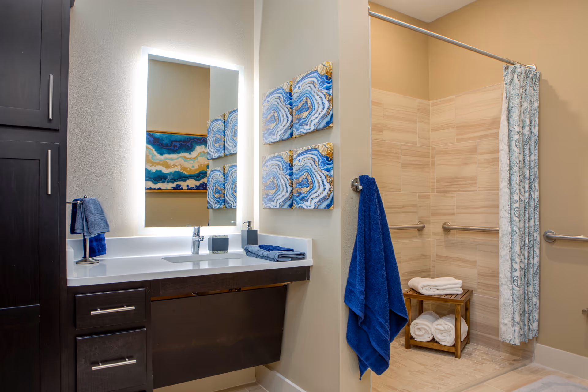 A modern bathroom featuring a vanity with a white countertop, a backlit mirror, and dark wood cabinets. To the right is a walk-in shower with beige tiles, a patterned shower curtain, grab bars, and a wooden stool holding folded white towels. Blue towels hang on a hook and a stand near the sink. Two abstract blue and gold artworks are mounted on the wall beside the shower.