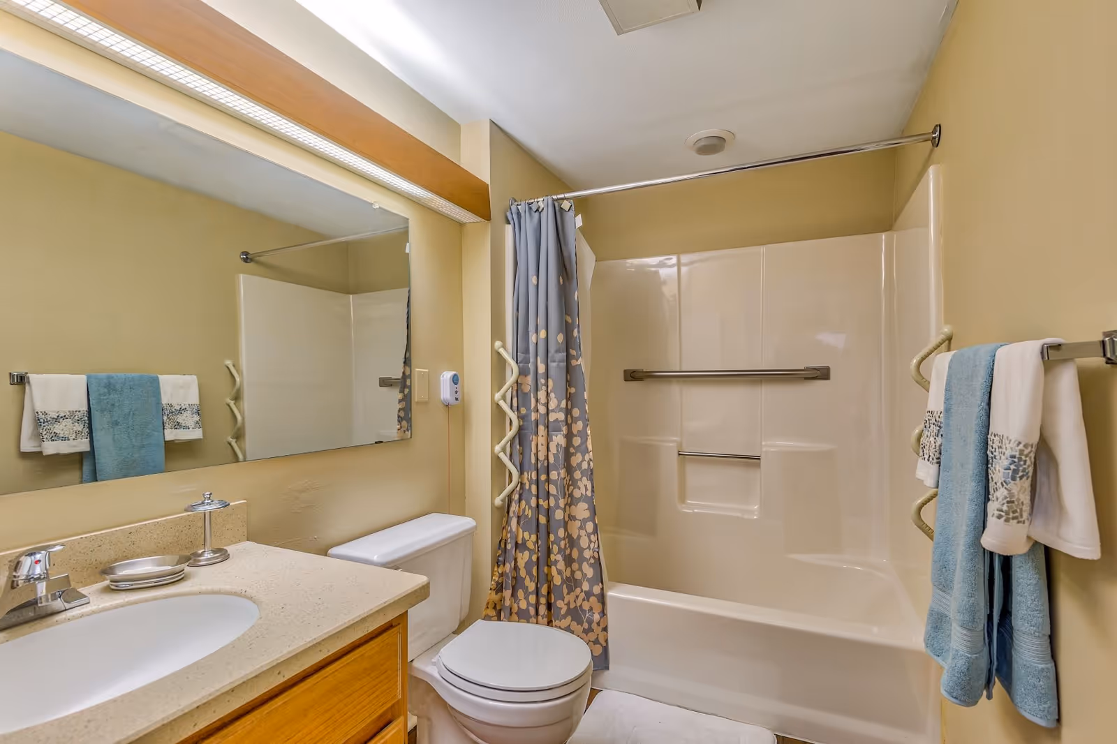 A clean bathroom featuring a beige countertop with a sink, a large mirror above the sink, a toilet, and a bathtub with a shower curtain decorated with a floral pattern. There are blue and white towels hanging on towel racks on the wall.