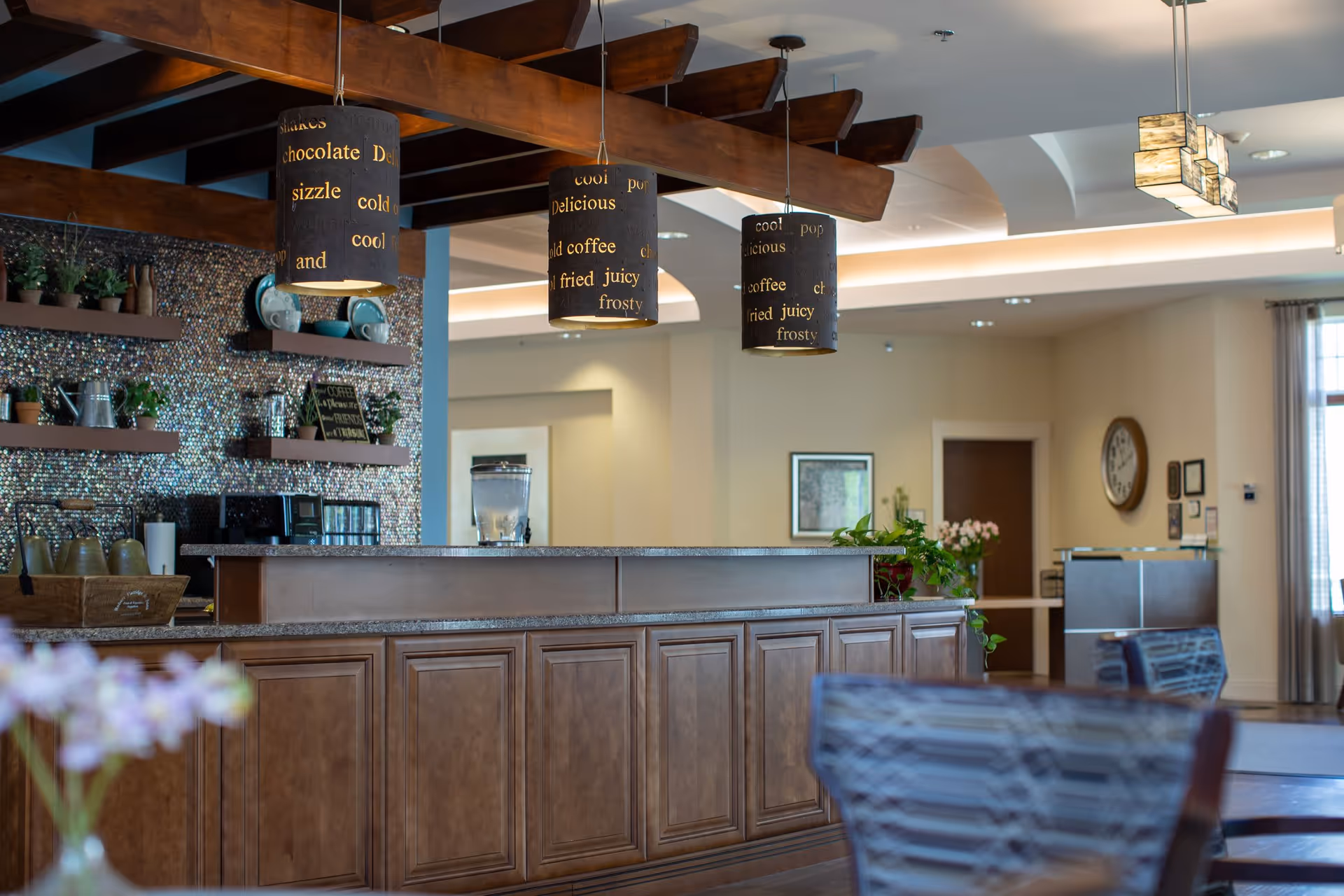 Interior view of a senior living facility's reception/cafe area with a wooden counter, hanging pendant lights, shelving with dishes and plants, and seating.