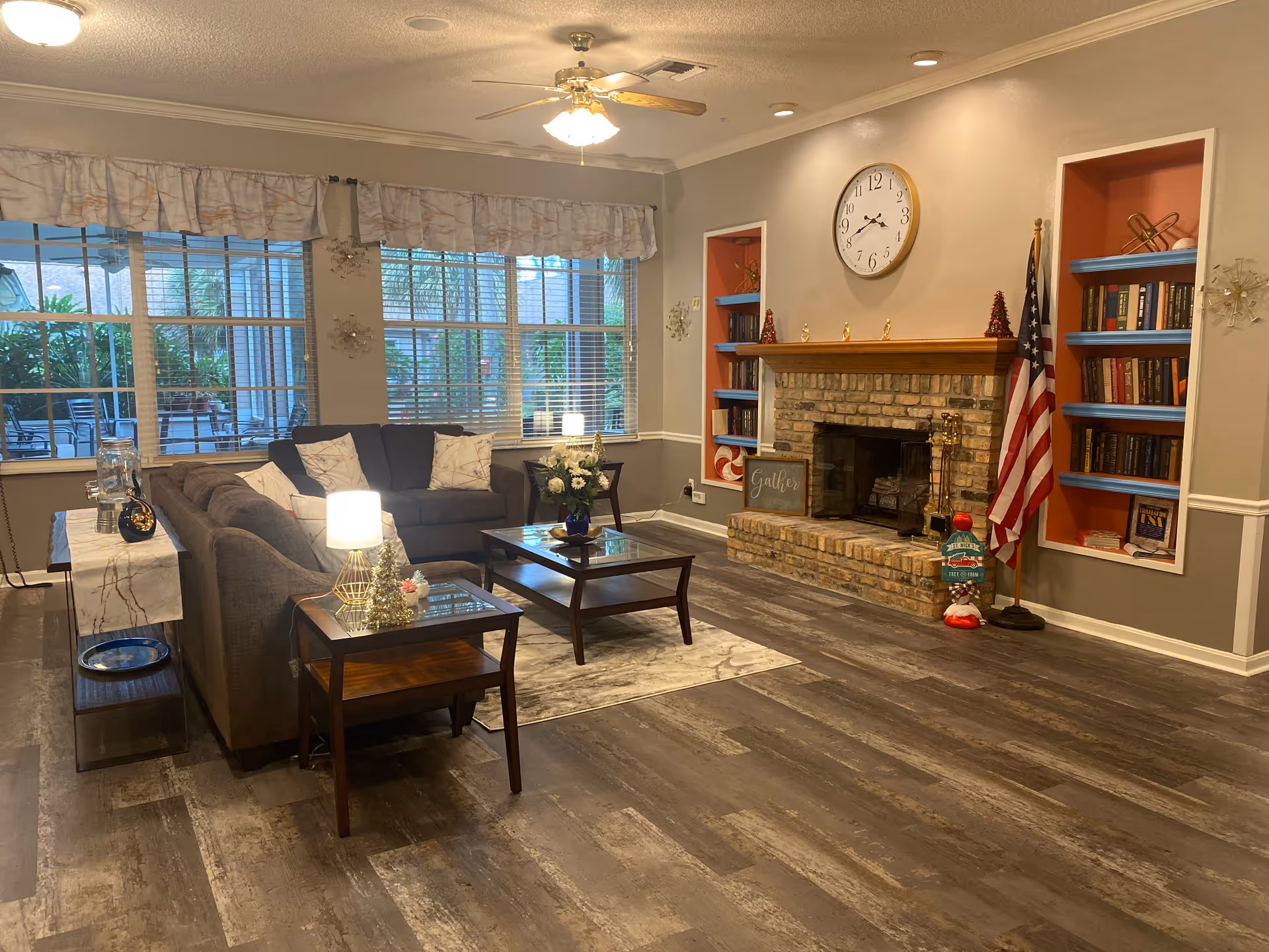 A cozy living room with a gray sectional sofa, two wooden coffee tables with glass tops, and a small lamp on a side table. The room features large windows with white blinds and valances, a brick fireplace with a wooden mantle, built-in shelves with books and decorative items, and an American flag standing next to the fireplace. The floor is covered with wood-look vinyl planks, and there is a ceiling fan with lights above.