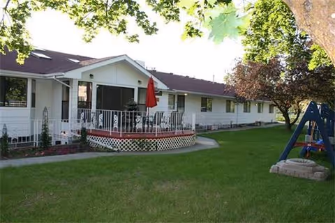 Single-story white senior living facility exterior with a green lawn, deck, and playground swing.