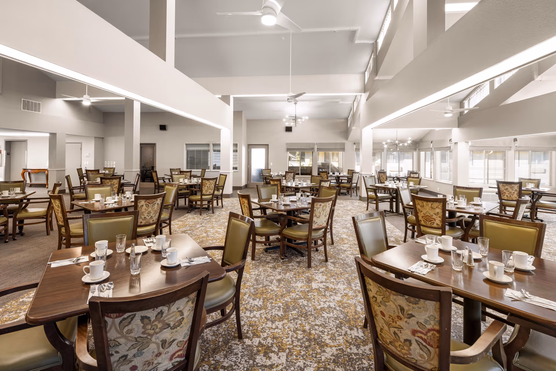 A spacious dining room with multiple wooden tables and chairs arranged neatly. Each table is set with white cups, glasses, silverware, and napkins. The room features high ceilings with ceiling fans and large windows allowing natural light to fill the space. The carpet has a patterned design, and the walls are painted in neutral tones.
