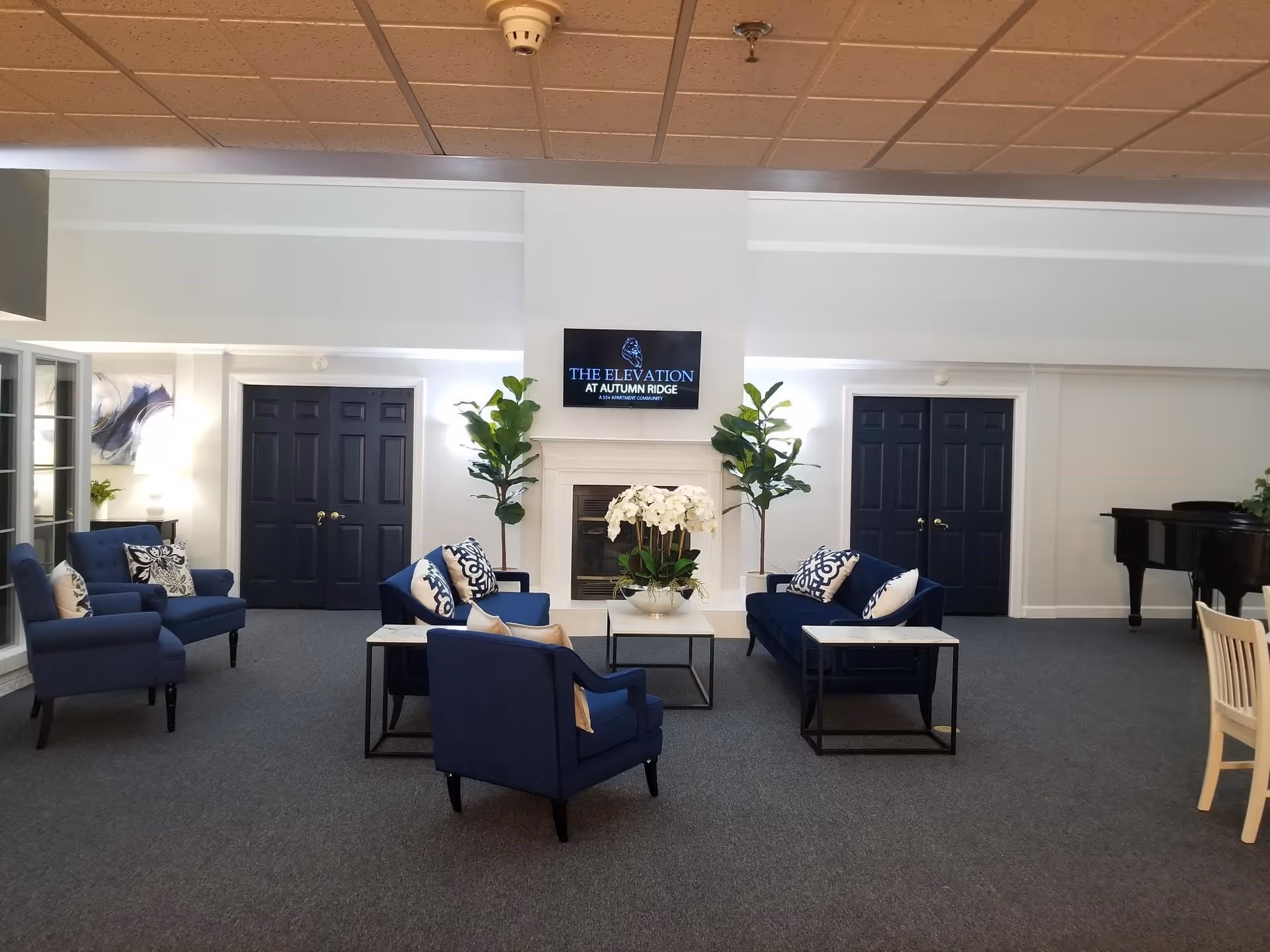 A spacious living room area with blue upholstered chairs and sofas arranged around a white coffee table with a white orchid centerpiece. There are two tall green plants flanking a white fireplace with a TV mounted above it displaying 'THE ELEVATION AT AUTUMN RIDGE'. The room has dark double doors on either side of the fireplace, a grand piano on the right, and a light-colored chair partially visible on the far right.