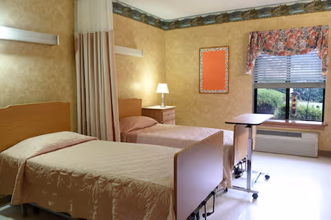 A room with two single beds covered with beige bedspreads, a bedside table with a lamp between them, a privacy curtain partially drawn, a window with floral valance and blinds, and a small overbed table on wheels near the window.
