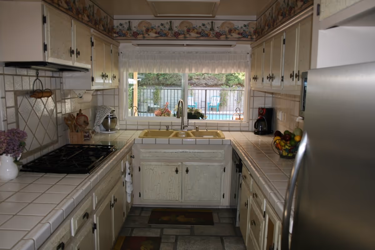 A kitchen with cream-colored cabinets and tiled countertops. There is a yellow double sink under a window with a white curtain, looking out to an outdoor area with a pool and patio chairs. The kitchen has a gas stove, a coffee maker, a stainless steel refrigerator, and a bowl of fruit on the counter.