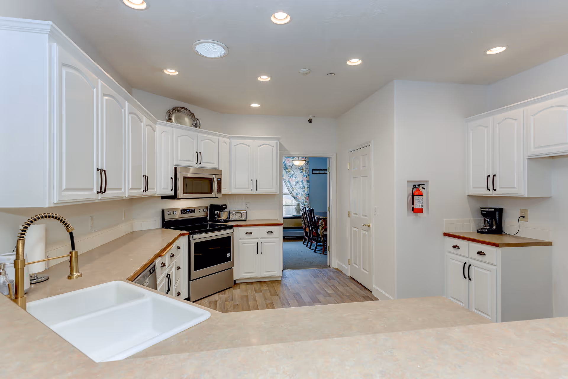 Bright kitchen with white cabinets, stainless steel appliances, a sink in the foreground, and a doorway leading to a dining area.