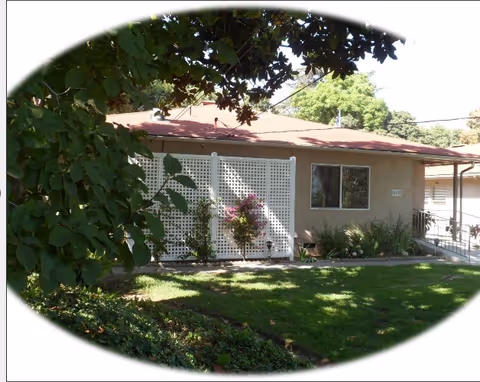 Single-story building with a red roof, beige walls, and a white lattice fence with plants growing in front. The building is surrounded by green grass, bushes, and trees providing shade.