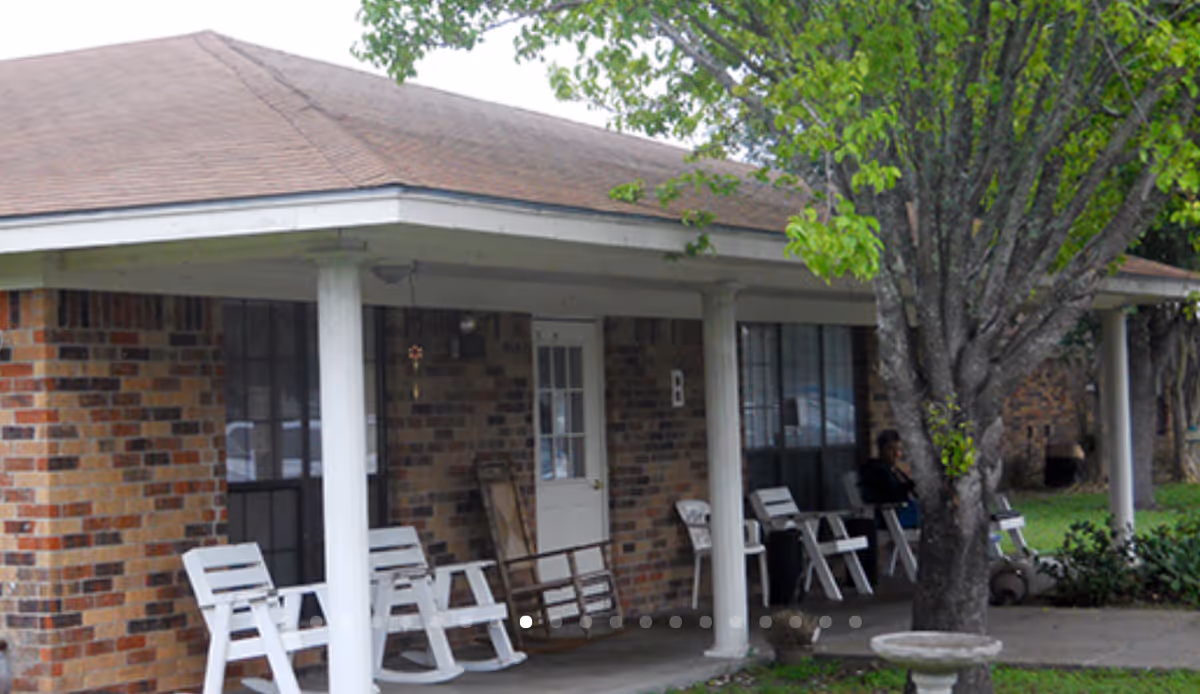 Covered front porch of a brick assisted living building with white rocking chairs and a tree in front.