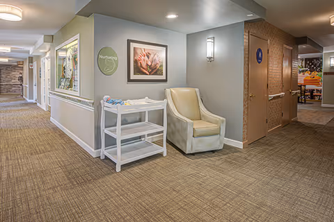 Carpeted interior hallway of a memory care facility with an armchair, a small white shelving cart, framed artwork, and doorways to other rooms.