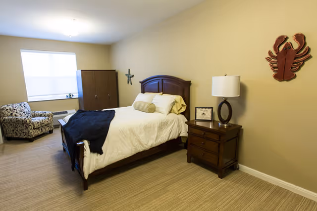 A neatly arranged bedroom with a wooden bed featuring white bedding and a dark throw blanket. There is a wooden nightstand with a lamp and a framed picture on it. On the wall above the nightstand, there is a decorative red lobster and a starfish. A patterned armchair is placed near a window with blinds, and a wooden wardrobe stands against the wall.