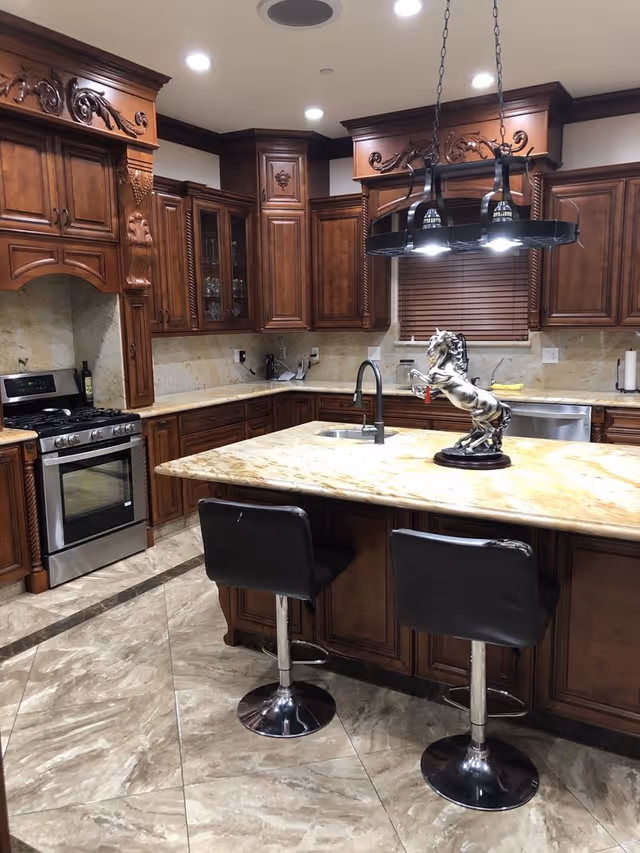 A kitchen with ornate wooden cabinets, a marble countertop island with two black bar stools, a stainless steel stove, and a decorative horse statue on the island. The kitchen has tiled floors and recessed ceiling lights.