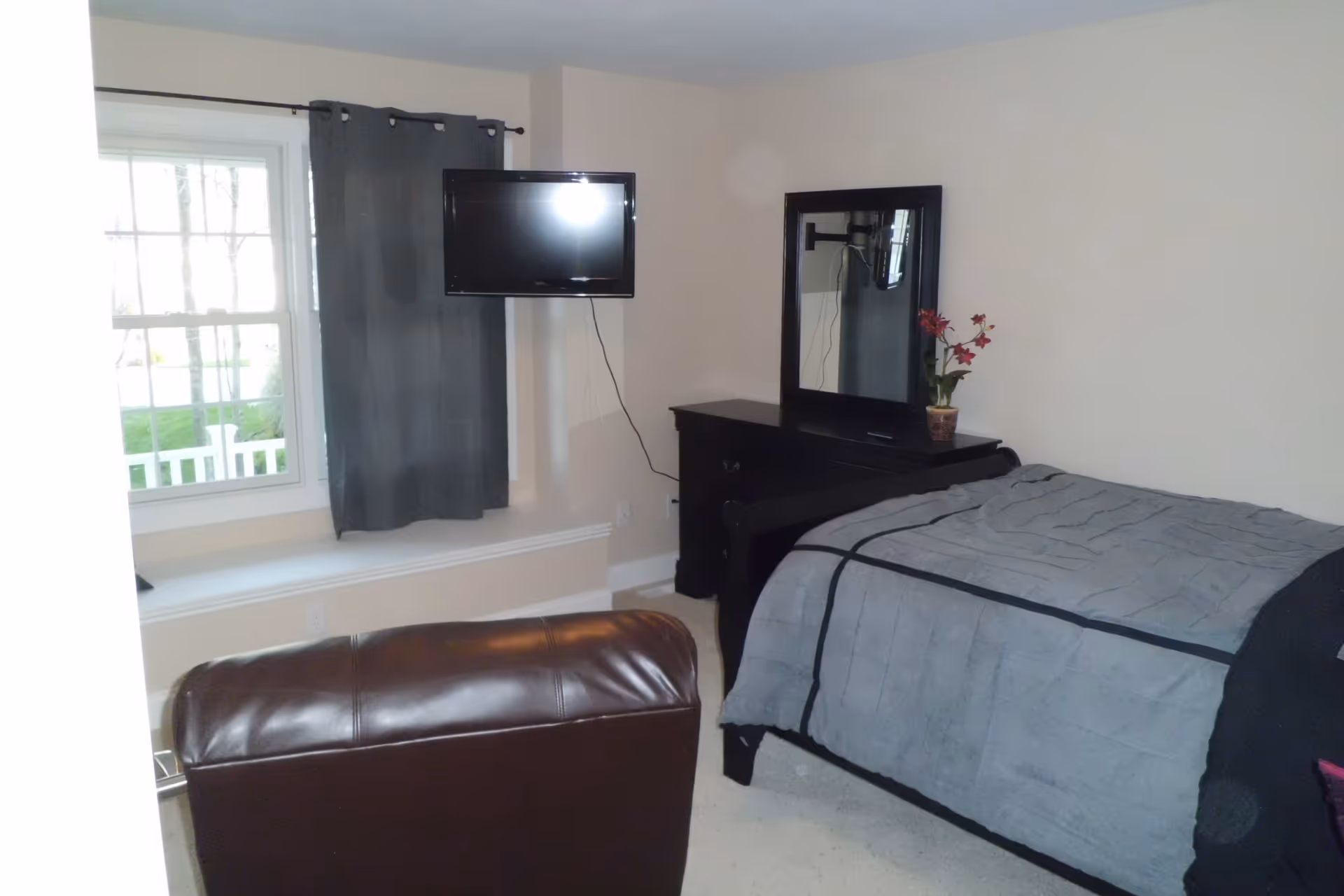 A bedroom with a bed covered in a gray blanket, a black dresser with a mirror and a potted plant on top, a wall-mounted flat screen TV, a window with gray curtains, and a brown leather chair in the foreground.