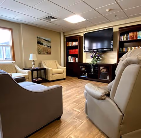 A cozy living room with beige and gray upholstered chairs and sofas arranged around a wooden floor. A dark wood entertainment center with shelves filled with books and a flat-screen TV mounted above is visible. A small side table with a lamp and a landscape painting on the wall add to the warm ambiance.