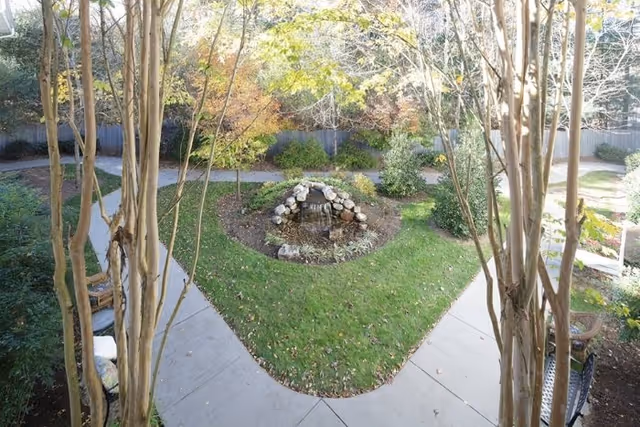 A landscaped outdoor garden area with a small rock waterfall feature in the center, surrounded by green grass, bushes, and trees. Concrete pathways form a Y shape around the garden, and there are benches along the paths.