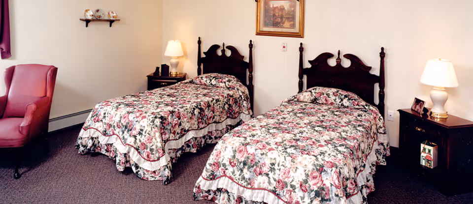 Two twin beds with floral bedspreads, wooden headboards, bedside tables with lamps, and a pink armchair in a senior living bedroom.