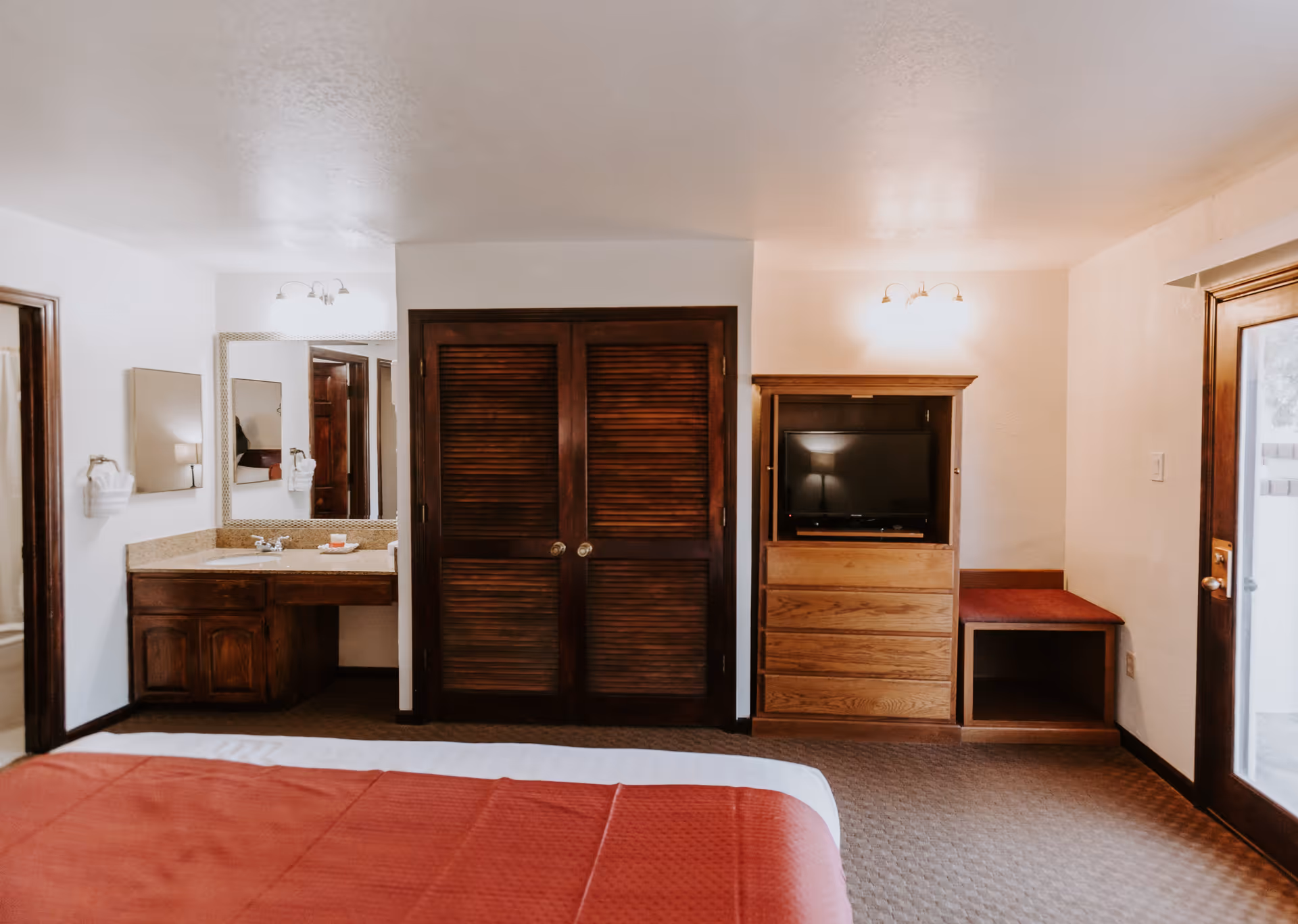 Interior view of a bedroom in Independent Living at Arizona Golf Resort featuring a bed with a red bedspread in the foreground, a wooden closet with louvered doors in the center, a wooden dresser with a TV on top to the right, and a small vanity area with a sink and mirror to the left. There is a door with glass panels on the far right side.