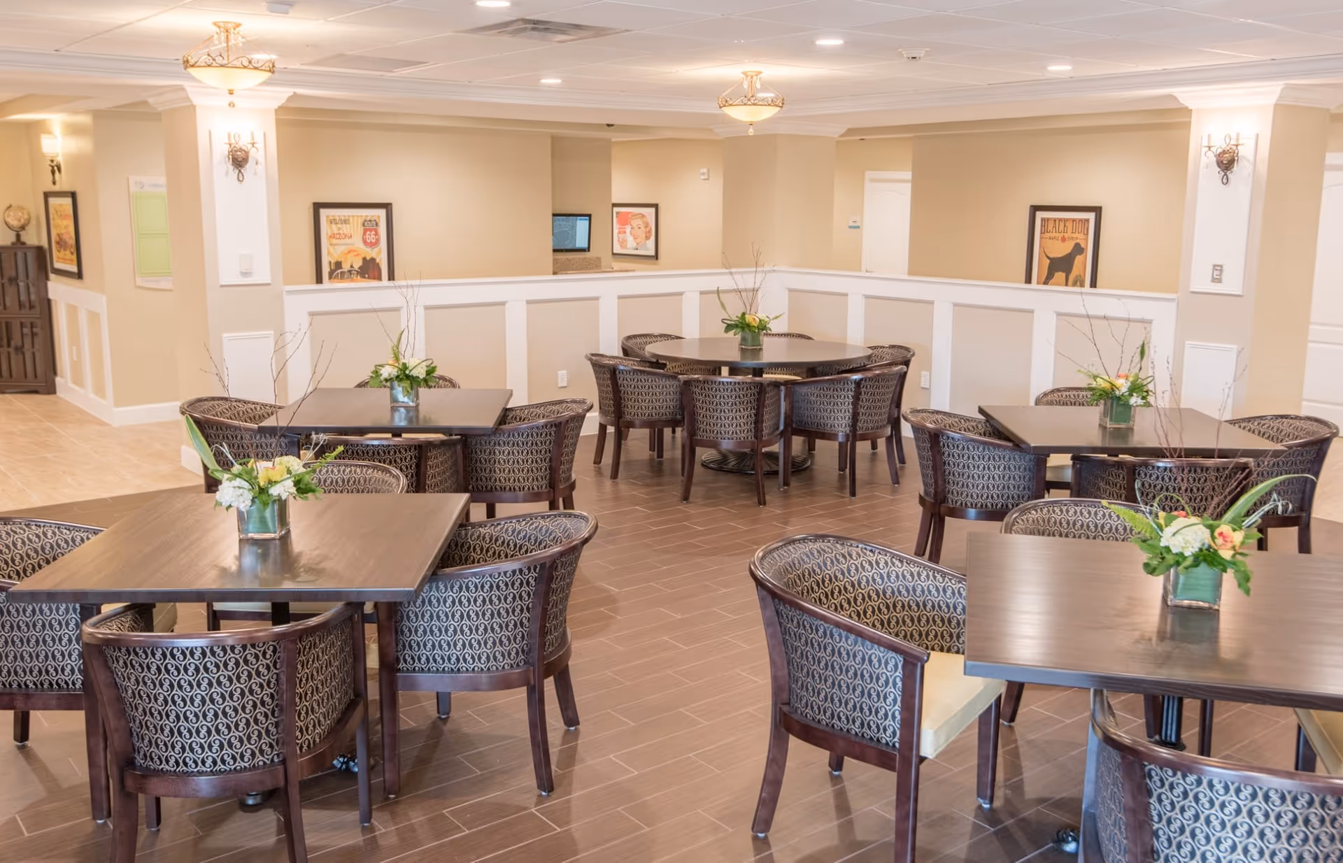 A bright and spacious dining area with multiple dark wood tables and patterned upholstered chairs arranged neatly. Each table has a small floral centerpiece. The room features beige walls with white wainscoting, framed artwork, and warm ceiling lights.
