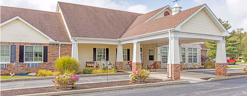 Front entrance of a single-story senior living facility showing a covered drive-through portico with columns, planters, and outdoor seating.
