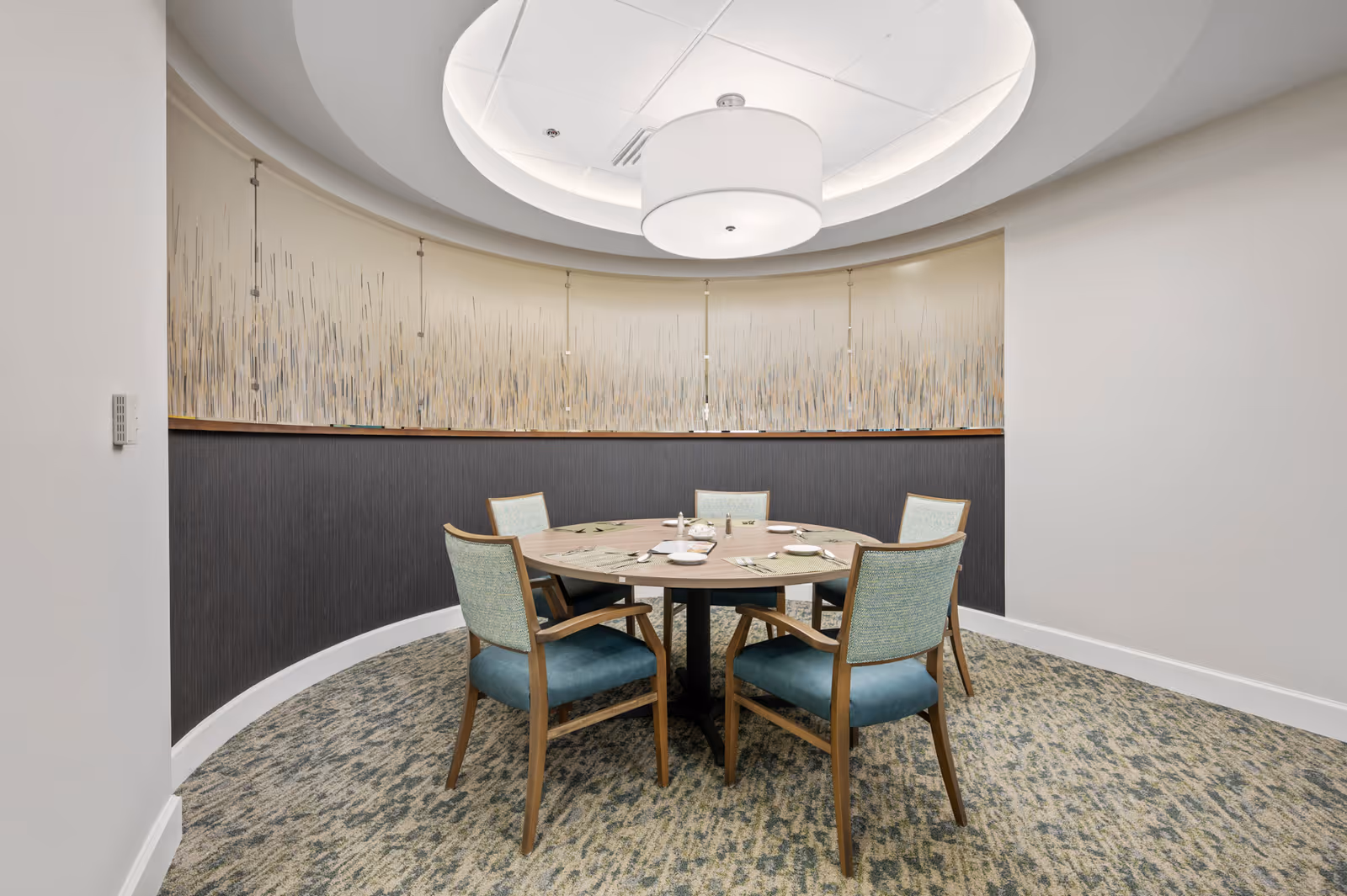 A small, round dining table set for four with plates, napkins, and utensils in a softly lit, circular room with patterned carpet and modern wall art. Four wooden chairs with teal cushions surround the table, and a large, round ceiling light fixture hangs above.