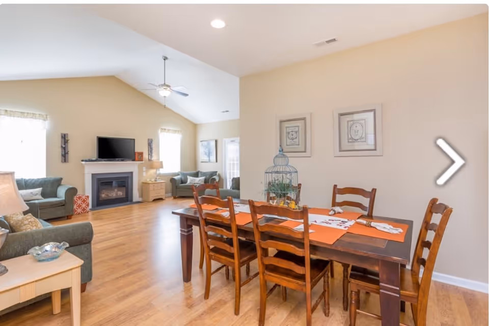 A bright and spacious living and dining area with wooden flooring. The dining area features a dark wooden table with six chairs and orange placemats. The living area has two green sofas, a fireplace with a mounted TV above it, and large windows letting in natural light. The walls are painted beige, and there are framed pictures hanging above the dining table.