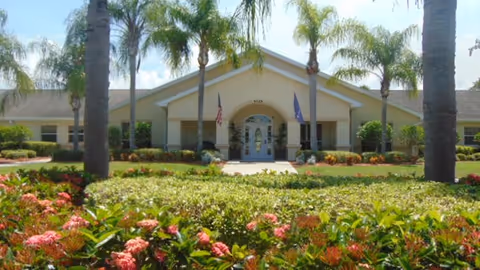 Front exterior view of Arden Courts - ProMedica Memory Care Community in Sarasota, showing a single-story building with a peaked roof, entrance with glass doors, two flags on either side, palm trees, and landscaped bushes and flowers in the foreground.