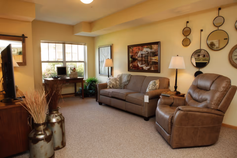 A cozy living room with a brown leather recliner and a brown fabric sofa with patterned pillows. There are two table lamps on either side of the sofa, a wall decorated with multiple round mirrors, and a framed picture of a rustic scene above the sofa. A wooden desk with a computer is positioned in front of a window letting in natural light. Two large decorative vases with dried plants are placed near a wooden TV stand with a flat-screen TV.