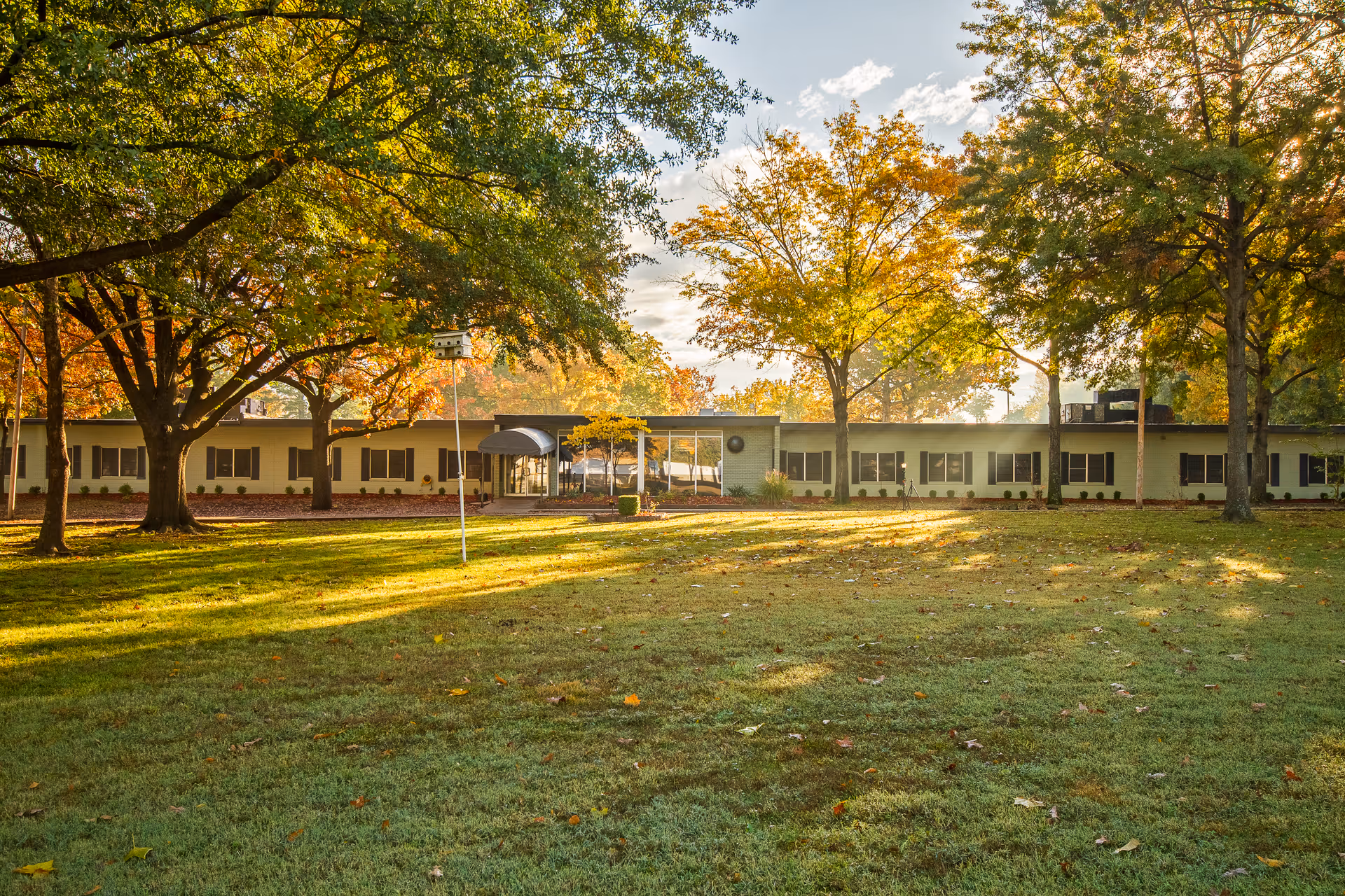 A single-story building surrounded by large trees with autumn foliage. The building has multiple windows and a covered entrance. The foreground features a grassy lawn with scattered fallen leaves, illuminated by sunlight filtering through the trees.